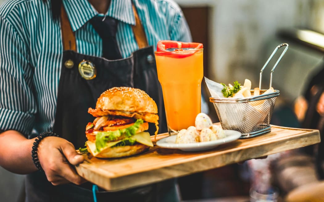 A person with an apron on carries a board of food including a drink, burger and fries.