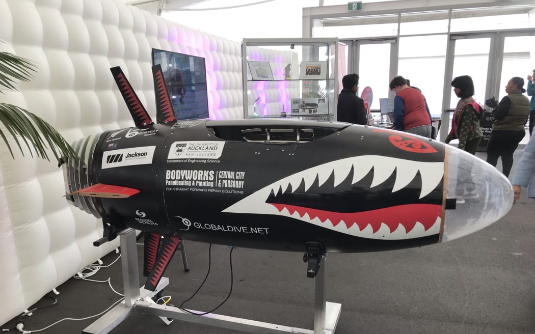 A torpedo shaped black machine sits on a silver metal stand, with sharp white teeth drawn on, and fin like things sticking out from the top and bottom.
