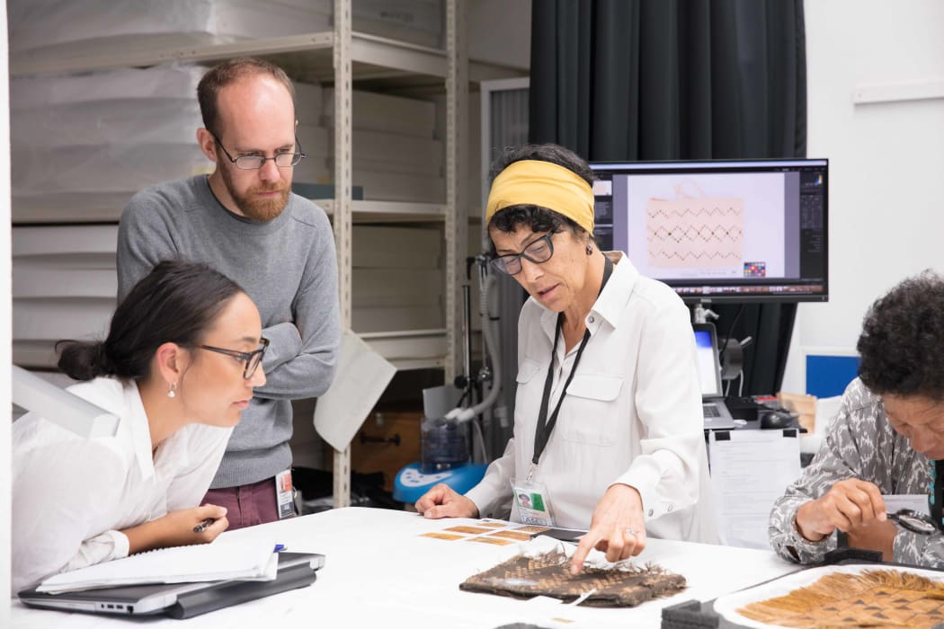Auckland Museum staff Jenna Dudley and Conor Tulloch discussing the storage and care of a kete muka (woven flax fibre bag) with textile conservator Rangi Te Kanawa.  Her sister Kahutoi Te Kanawa is inspecting another kete muka on the table.