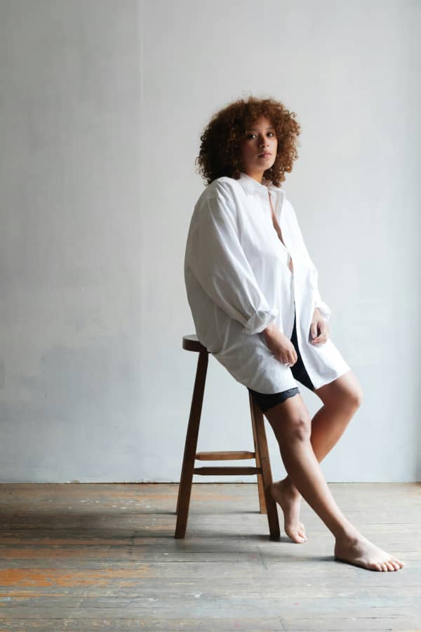 A woman with a red afro sits on a stool wearing a white shirt.