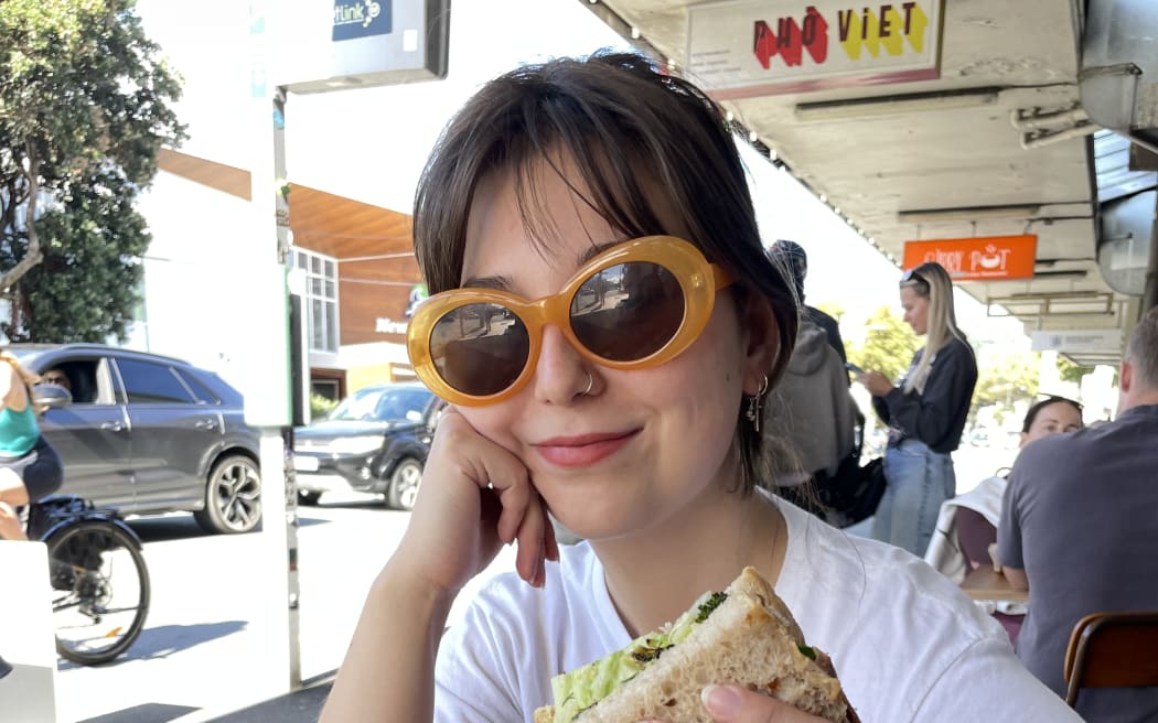 Bryer is sitting at a cafe table on a busy Wellington street. She smiles at the camera, wearing funky yellow sunglasses and holding half of a sandwich.