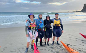 Four men on a beach, standing in a line, pose for the camera, smiling with their arms around each other.