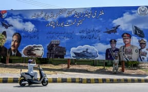 A man clicks a picture of a billboard featuring Pakistan's Army Chief General Syed Asim Munir (C), Naval Chief Admiral Naveed Ashraf (R), and Air Chief Marshal Zaheer Ahmed Babar Sidhu, along a road in Peshawar