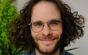 Mid-shot of NPR Podcast host Darian Woods who has long curly hair and wears glasses