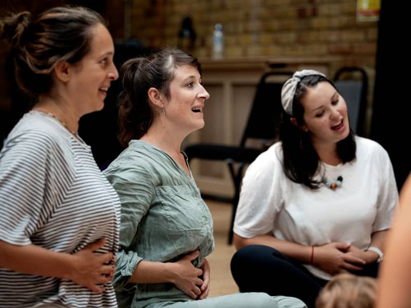 Three mums place their hands on their stomachs as they sing.