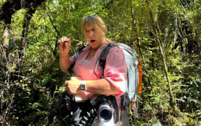 This is a photo of Paula Vigus in a forest. Her cameras are dangling around her waist. She is holding a twig with a fungi on it and looking at it in surprise.
