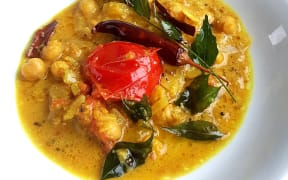 A close-up image of coconut chickpea curry on a shallow white bowl, decorated with curry leaves and a tomato.