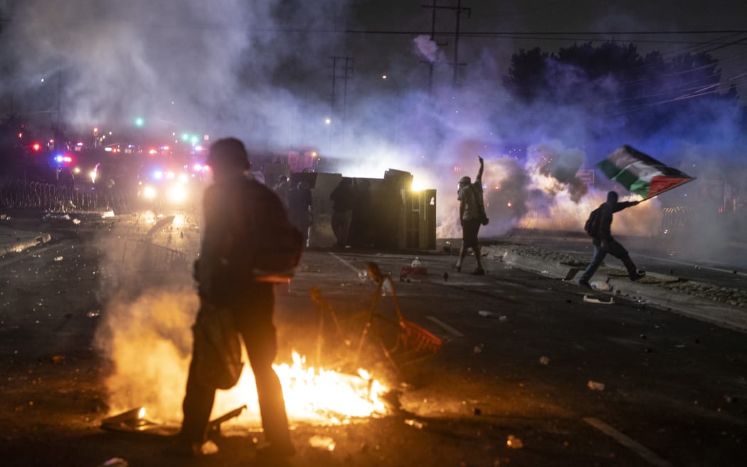 A protester waves the Palestinian flag (R) as law enforcement clashes with demonstrators during a protest following federal immigration operations, in the Compton neighborhood of Los Angeles, California on June 7, 2025. US President Donald Trump deployed 2,000 troops on June 7, 2025 to handle escalating protests against immigration enforcement raids in the Los Angeles area, a move the state's governor termed "purposefully inflammatory." Federal agents clashed with angry crowds in a Los Angeles suburb as protests stretched into a second night Saturday, shooting flash-bang grenades and shutting part of a freeway amid raids on undocumented migrants, reports said. (Photo by RINGO CHIU / AFP)