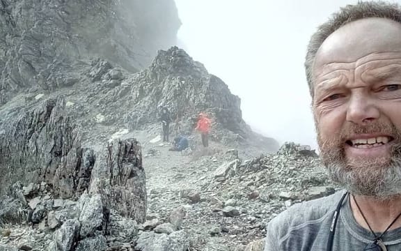 High up in Waiau Pass, Bruce pauses for a selfie in the background, two other walkers are climbing a steep, rocky ridge.