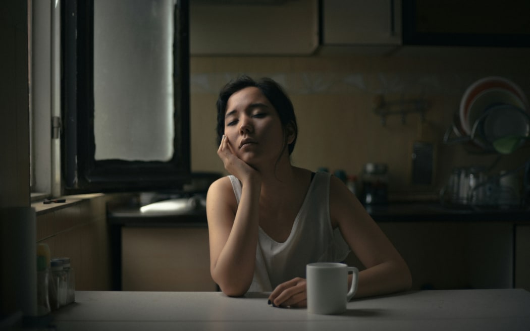 A woman sits thoughtfully at a kitchen table with a mug.
