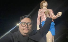 A man in a coat holds up a freshly cooked bacon sandwich up to a giant statue of a kid in blue shorts and a red and white striped shirt.