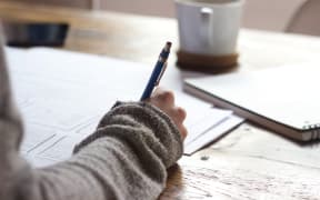 A hand holding a pen hovers over paper with a mug in the background.