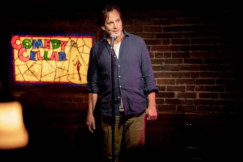 Will Arnett, wearing a partially buttoned blue shirt, talks into a standing microphone with the words 'Comedy Cellar' on an illuminated sign behind him.