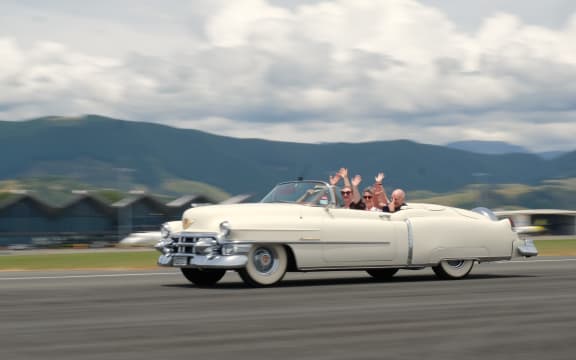 Fifties car drives down runway with four people  waving