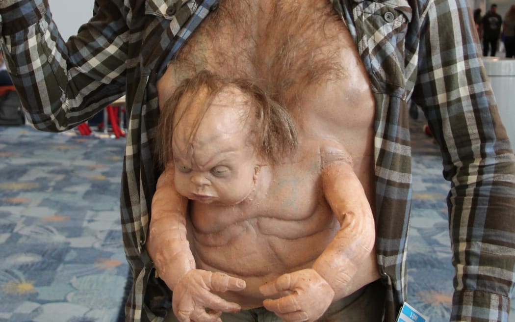 A man in cosplay of Kuato from Total Recall, so he has a baby-like doll coming out of his stomach and holding a gun above his head.