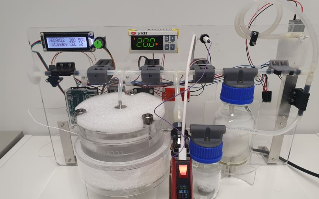 A complex scientific device on a white lab bench, with sensors, transparent chemicals in flasks, tubes and wires.