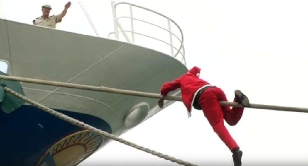 Halfway up the mooring rope, a Santa is refused entry to the ship by a uniformed guard.