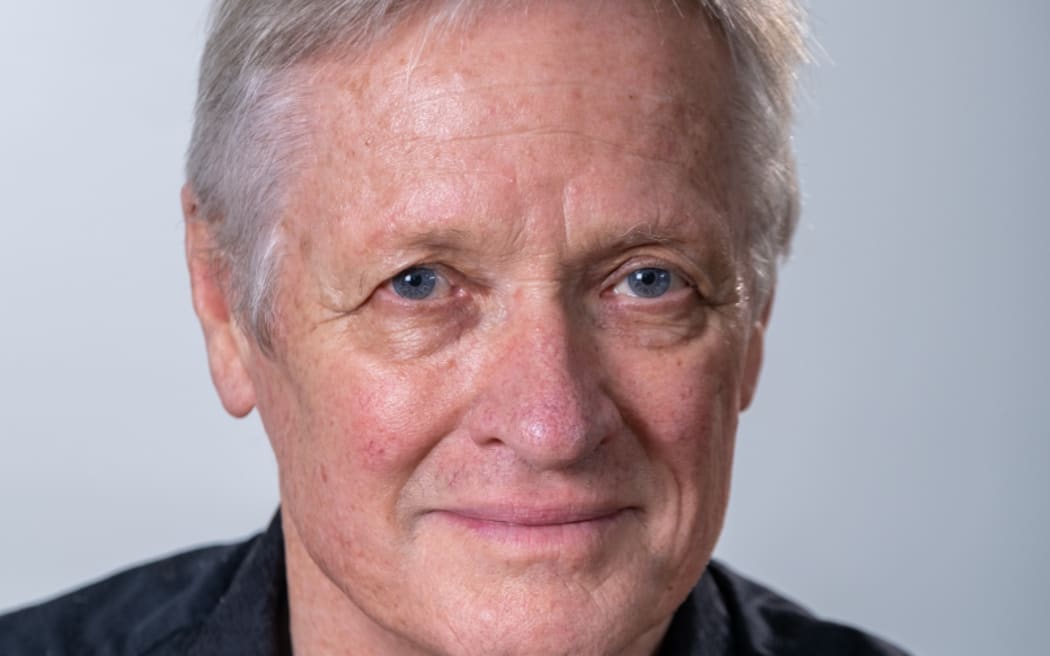 Opera director John Davies looks at the camera. He stands in front of a grey background. John looks like he's in his late 50s. His main feature is piercing blue eyes that almost feel as though they are looking through the camera and into your soul.