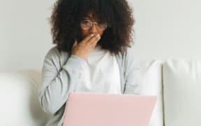 A woman wearing a white tshirt and grey cardigan covers her mouth with her hand in surprise as she looks at a laptop.ggen