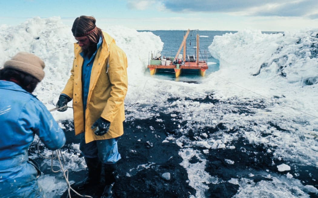 At the bottom left of the image, two people dressed in warm coats and gloves handle a length of rope. They are standing on frozen, rocky ground. Behind them is a small, industrial-looking boat parked in a small alcove in the sea, which has been carved out of large piles of ice.