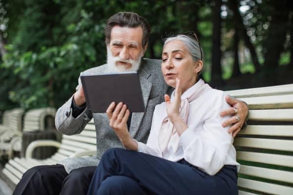 An elderly couple sit on a park bench speaking into an iPad.