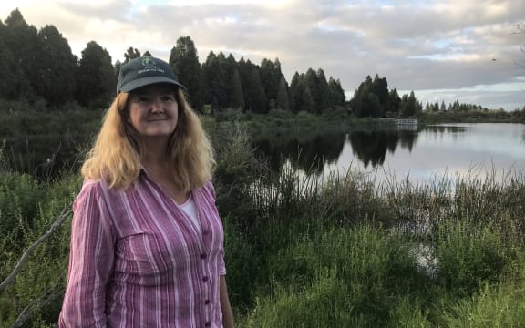 A woman wearing a cap stands in front of a lake and wetland vegetation on a cloudy day.