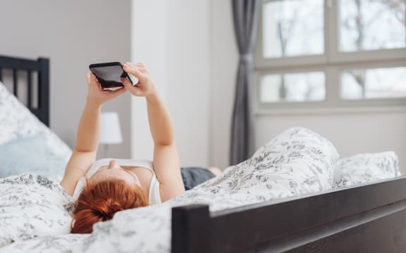Young woman spending a relaxing day at home lying on her back on the bed reading a message on her mobile phone