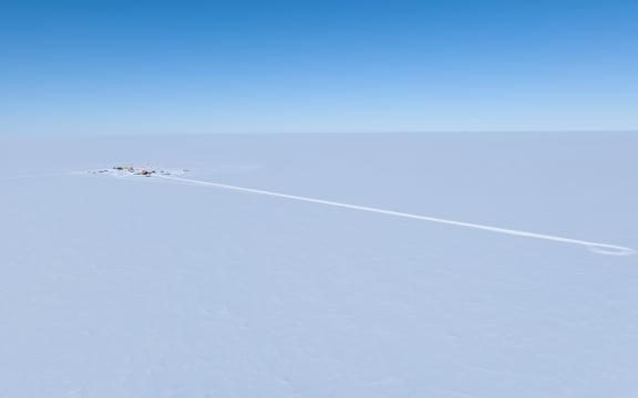 A drone image of the white expanse of flat ice beneath a clear blue sky with a tiny camp off to the left, and a long trail in the ice extending to the right.