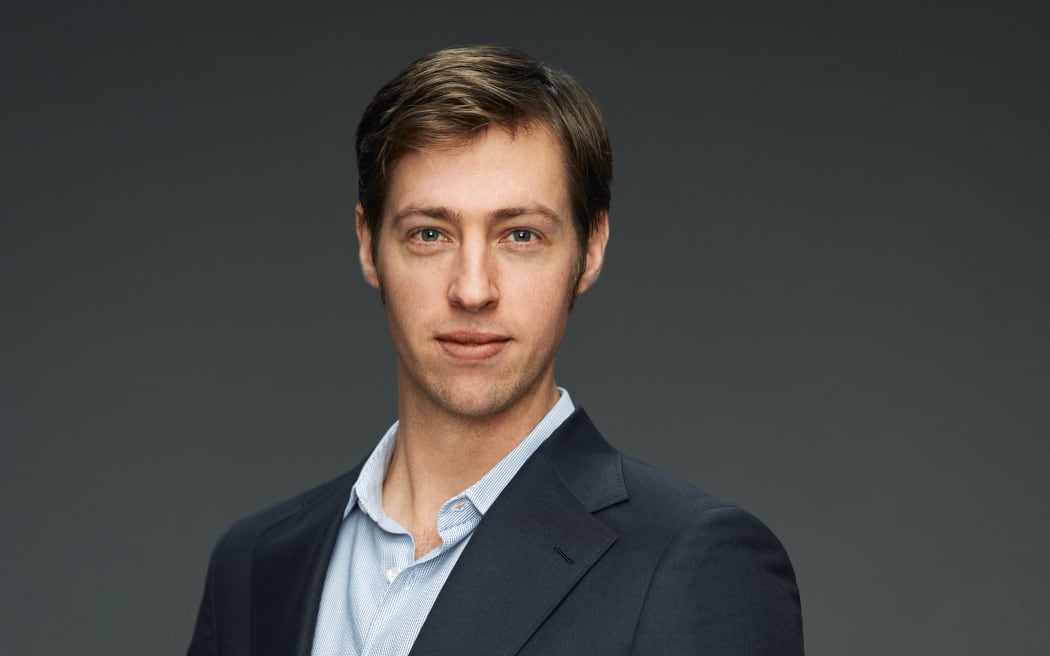 Ty King-Wall is stood in front of a grey background. He is looking directly at the camera and wearing a blue shirt and black jacket. Presumably he is wearing the matching suit trousers but they can't be seen in the shot. He has a very piercing gaze that gives the impression he's really looking deeply at you, but in a friendly way.