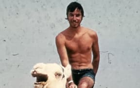 Chris Watson is riding a camel through the Sahara desert. He is shirtless. The sand behind him is very flat.
