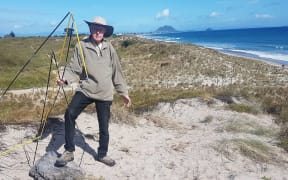 Pāpāmoa Beach resident Bruce Hutchinson stands in a bare patch of damaged and trampled dunes and holds some of the rubbish left behind following the party on 26 November, 2022.
