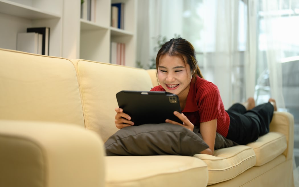 Happy young woman enjoying content on her digital tablet, lying on a sofa at home.