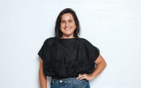 Sophie is standing against an off white background. It shows her from the waist up. She is wearing blue jeans and a black top tucked in at the waist. She had her left hand on her hip in a kind of classic 90s pose.