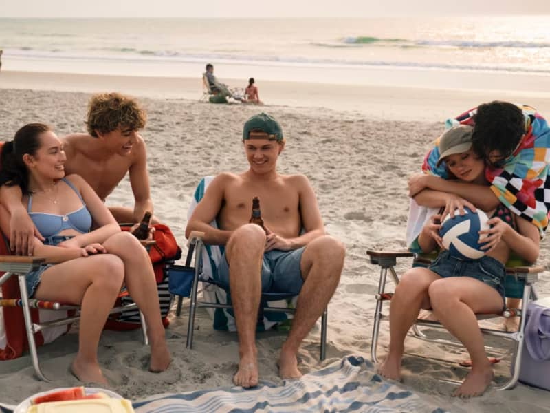 Five young people relax on a beach in deck chairs.