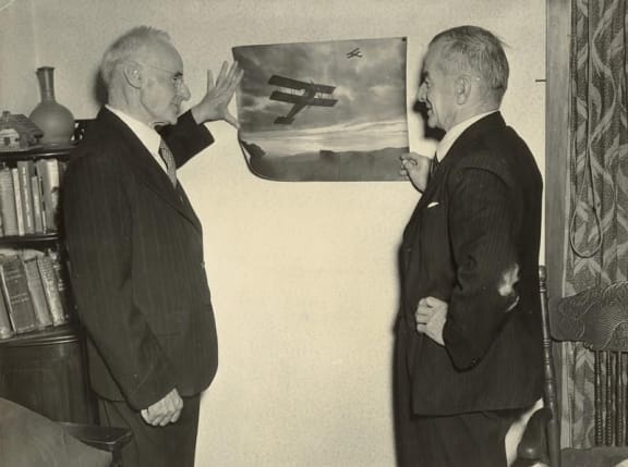 Image Leo and Vivian Walsh, 1940s, standing in front of a picture of  one of  their earliest aeroplane.