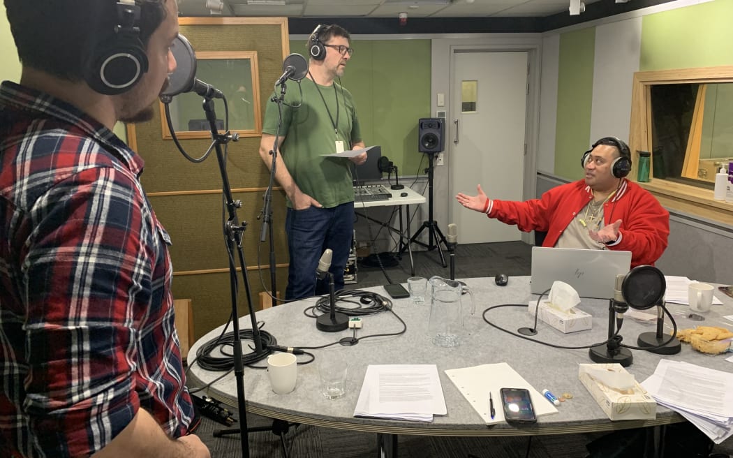 A photo of Co-hosts, James Nokise and John Daniell in the process of recording the podcast. They are in the studio standing at their mics. Seated at  a table is the language programme director, Matt Tufuga.