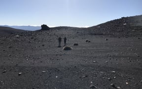Two people holding equipment stand in the mid ground. The scenery is dark gravel and rocks, it looks very much like a moonscape, with a smattering of plants. There's a clear blue sky in the background.