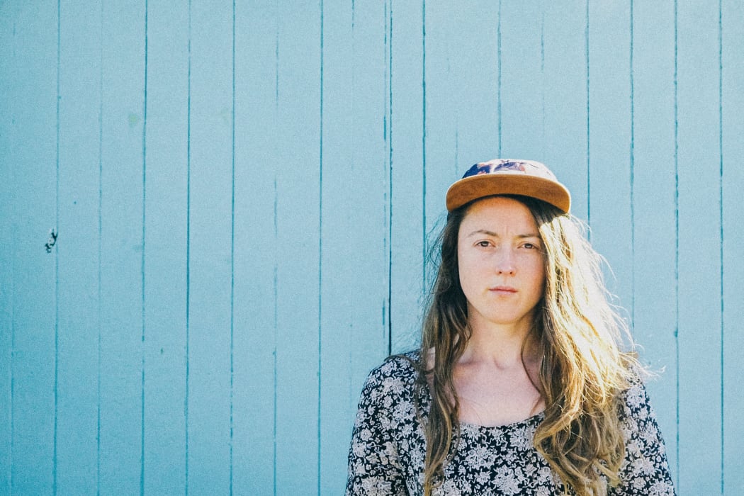 Person with long hair and cap standing in front of a blue painted wall