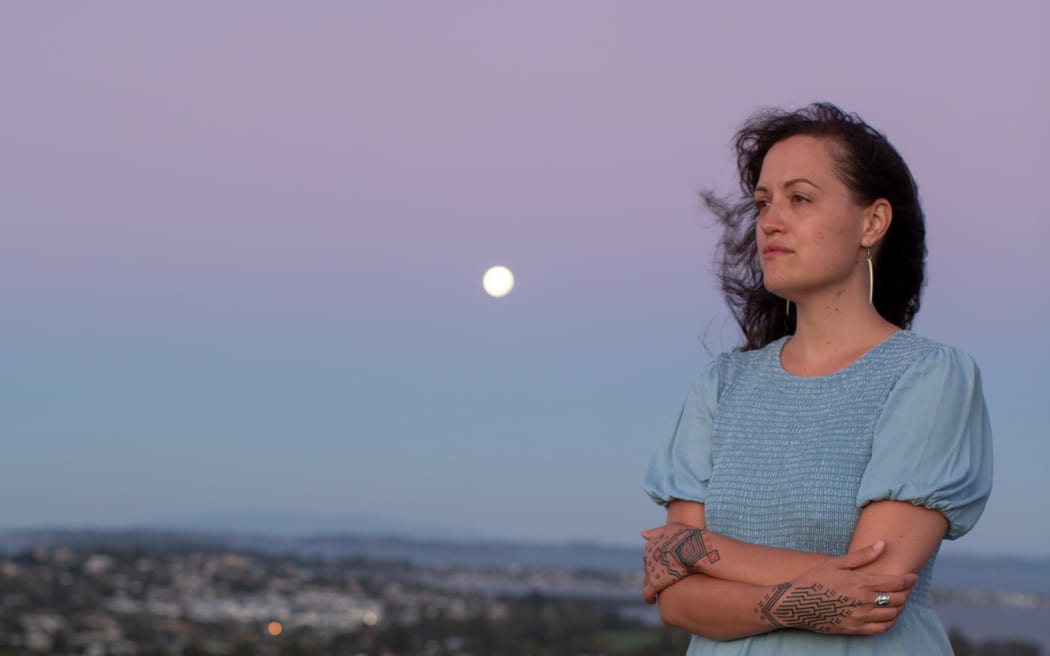 Māori architect Jade Kake wears a blue dress and stands in front of a city scape at sunset with her arms crossed in front of her. The moon can be seen in the background.