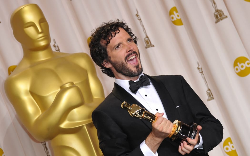 Bret McKenzie makes a zany face and holds an Academy Award, as he stands in front of a giant gold Oscar statuette.