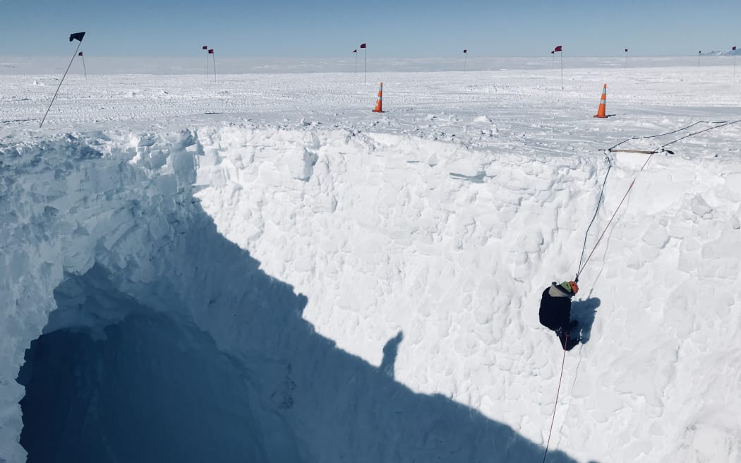 A person abseiling down the side of an icy crevasse. A flat white expanse of ice extends as far as the eye can see. There are two orange road cones visible along the edge of the crevasse, and a line of flags on poles further in the distance.The sky is hazy and blue.