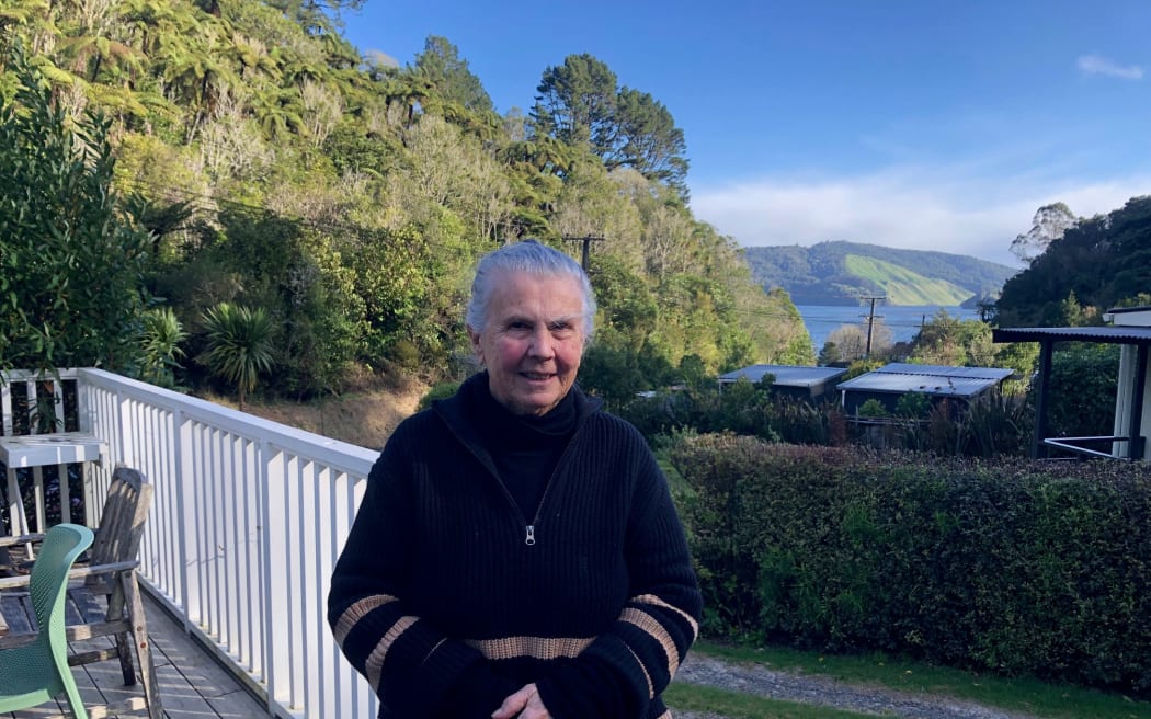 Portrait of an older woman, who is standing on the deck of her home. It is surrounded by bush and there is a glimpse of the nearby lake.