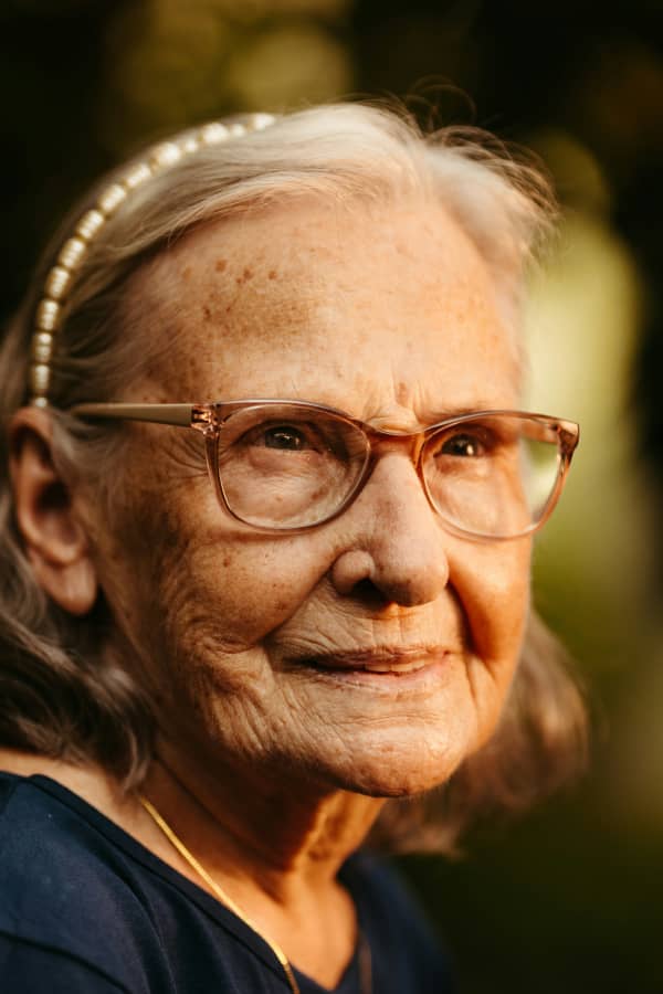 A senior woman smiles slightly and wears brown glasses.