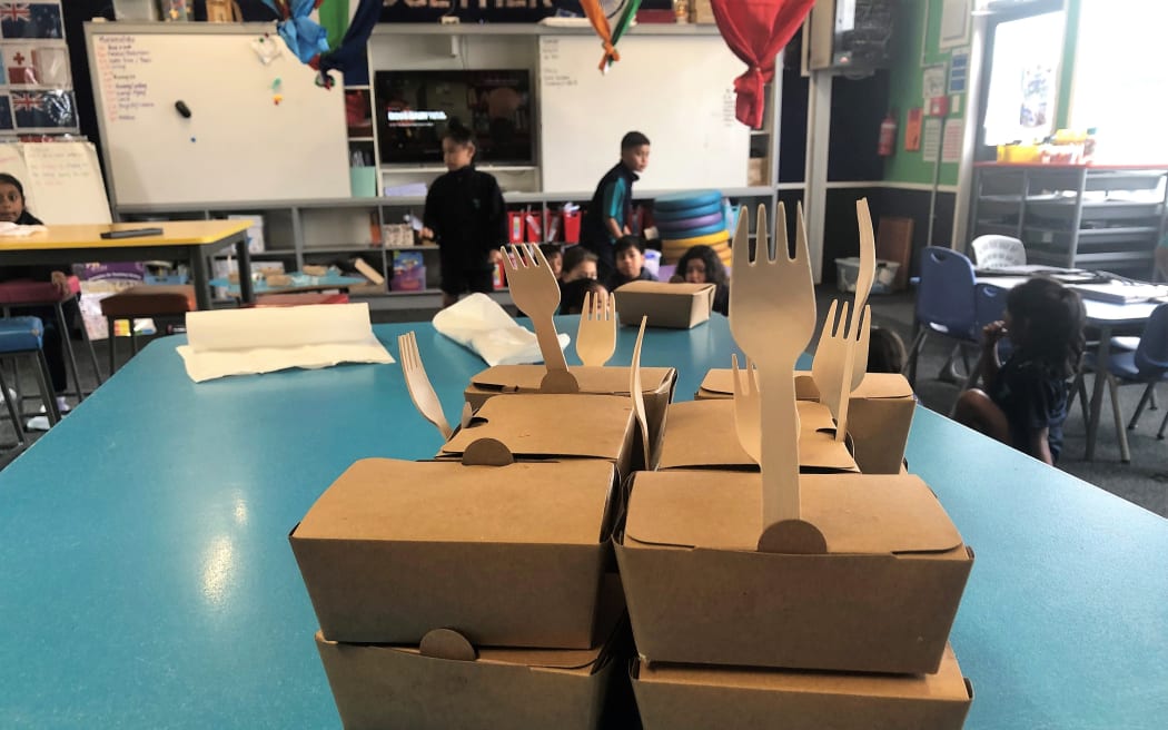 A stack of brown cardpaper boxes holding school lunches sit on a blue school table, with wooden forks sticking out of them.