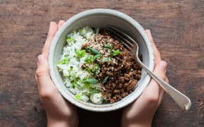 A bowl of stir-fried beef mince and rice garnished with spring onions and sesame seeds, held between two hands on a wood-grained surface.