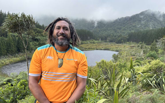 Mohi is standing at a point overlooking the Killarney lakes. The bigger lake is visible behind him with pine forest behind it. There's a lancewood tree directly behind him, and some harakeke and native trees visible around him. He's smiling, wearing a bright orange t-shirt. Both his hands are clasped together in front of him.