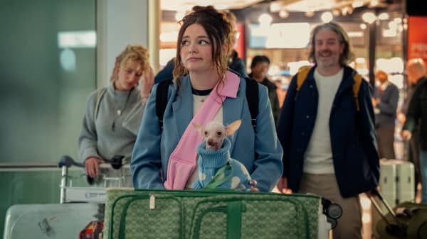 Meg Stalter (as Jessica in the Netflix series Too Much) at an airport pushing a trolley with luggage and a chihuahua on top.