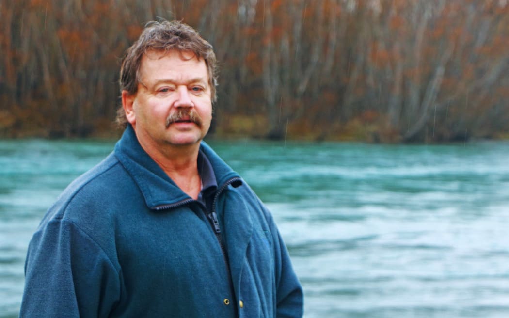 Man with moustache in fleece jacket stands in front of wide river