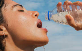 A woman opens her mouth to drink from a water bottle in the sun outdoors.