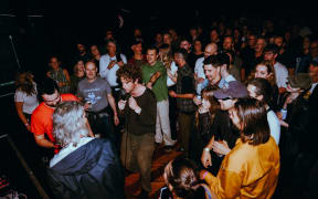 People crowd around a brown-haired male singer who stands among the audience at a live music gig.